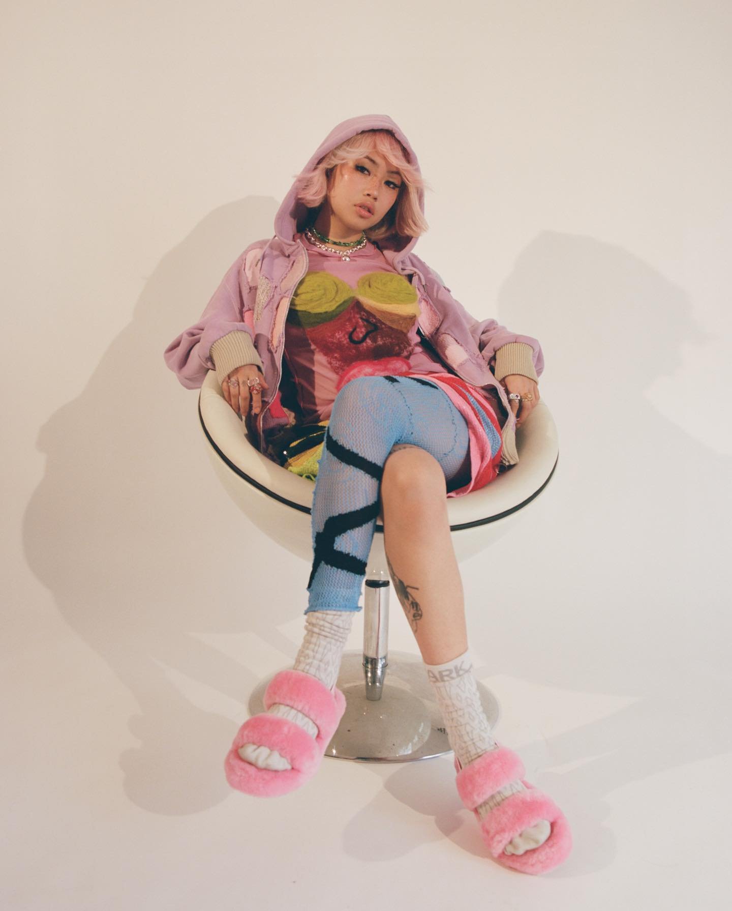 Young woman with pink hair sits in a modern chair wearing a colorful outfit and pink fuzzy slippers in a neutral studio setting.