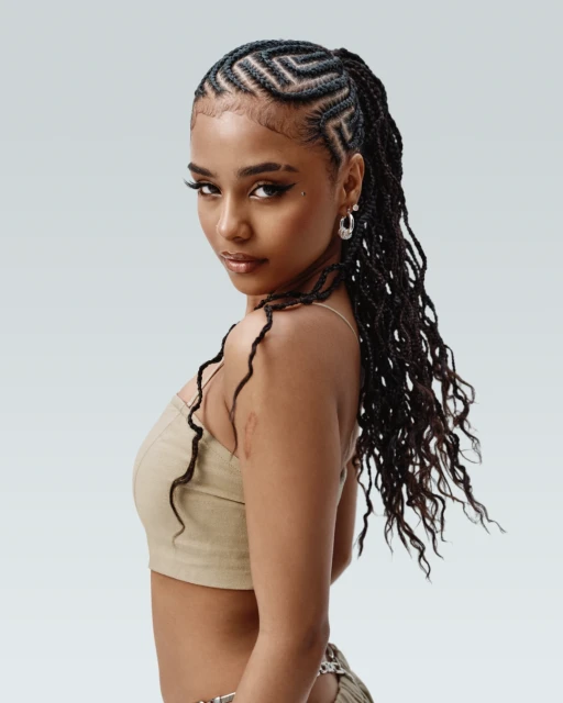 Portrait of a young woman with braided hair, wearing a neutral-toned crop top, posing in front of a light grey backdrop in a studio setting.
