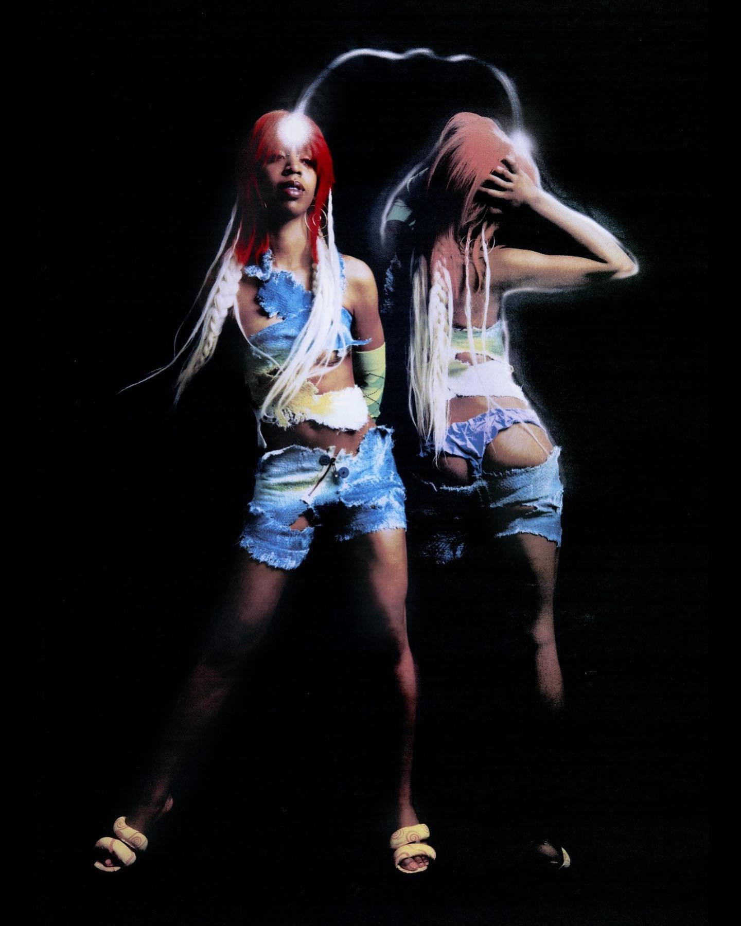 Two women with colorful hair and deconstructed outfits pose in a studio, lit by a soft glow against a dark backdrop, showing fashion and style.