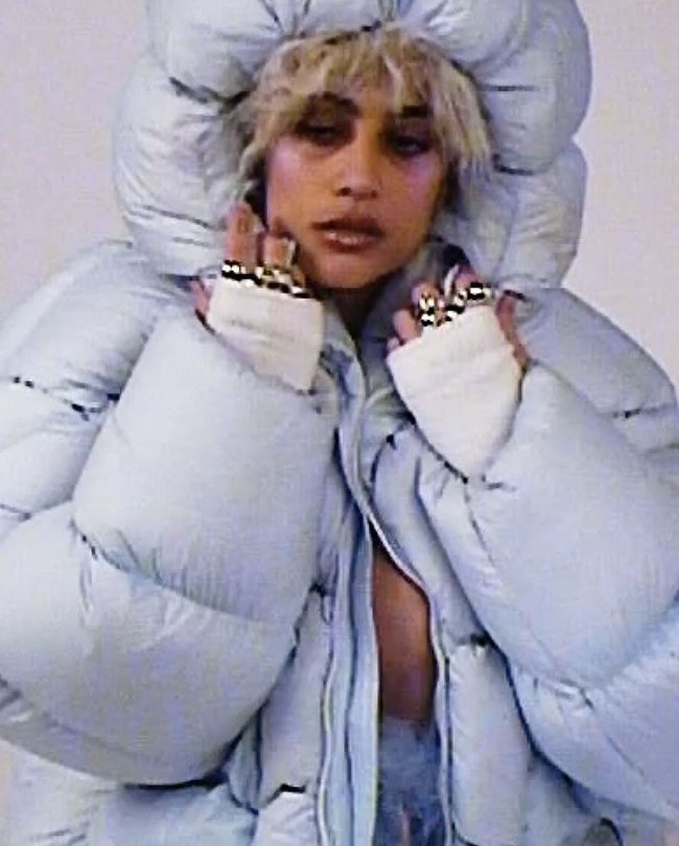 Young woman in a puffy hooded jacket poses in a photo studio with her hands up, displaying rings on her fingers and light makeup.