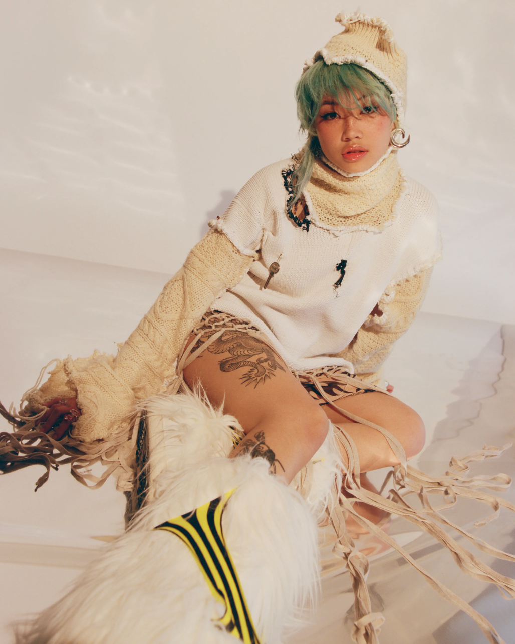 A young adult woman with green hair wears a knit cap and sweater indoors, posing with an artistic, avant-garde fashion style in soft light.