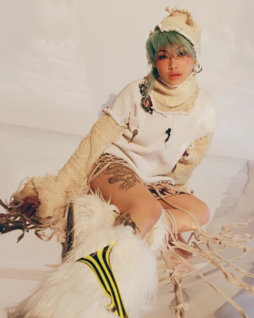 A young adult woman with green hair wears a knit cap and sweater indoors, posing with an artistic, avant-garde fashion style in soft light.