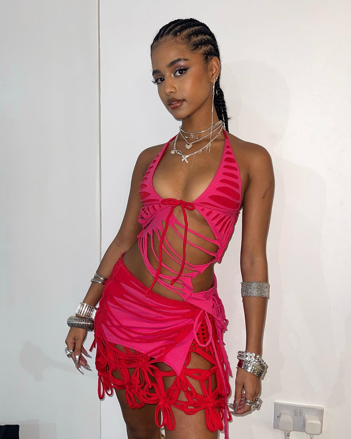 Young woman poses indoors wearing a pink and red halter top and skirt with braided hair, silver jewelry against a white background.