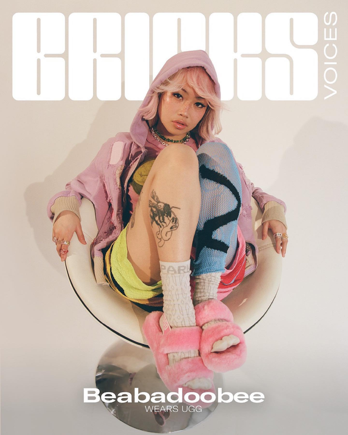 Beabadoobee poses on a white chair wearing UGG slippers, a pastel hooded jacket, and colorful leggings in a studio setting.