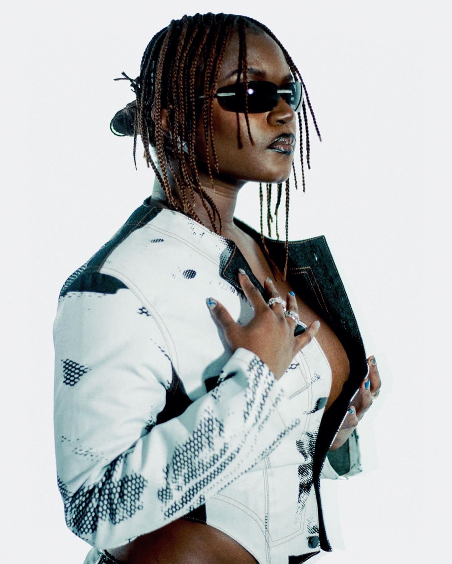 Stylish woman with box braids and sunglasses poses in a white jacket against a bright backdrop in a fashion-forward studio portrait.
