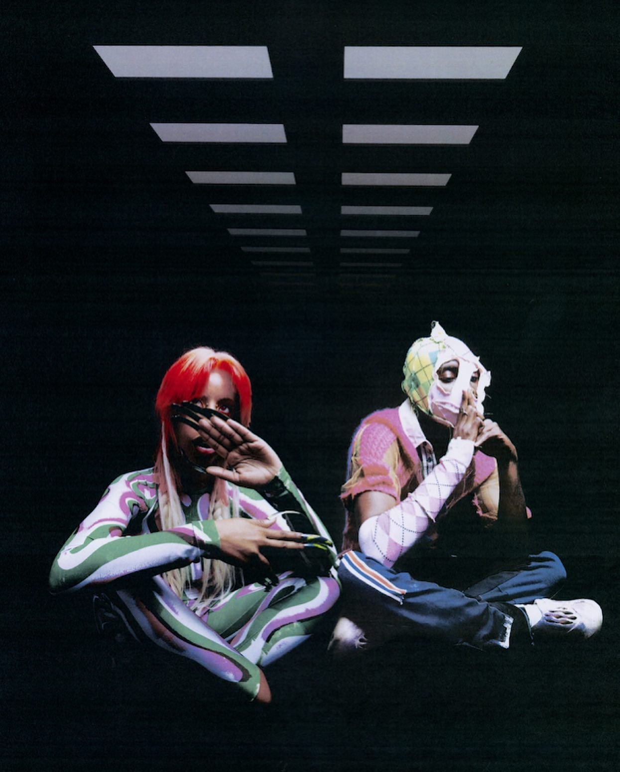 Two people sit in patterned outfits against a dark background with white ceiling lights creating a visually striking and surreal composition.