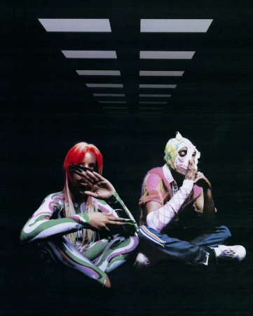 Two people sit in patterned outfits against a dark background with white ceiling lights creating a visually striking and surreal composition.