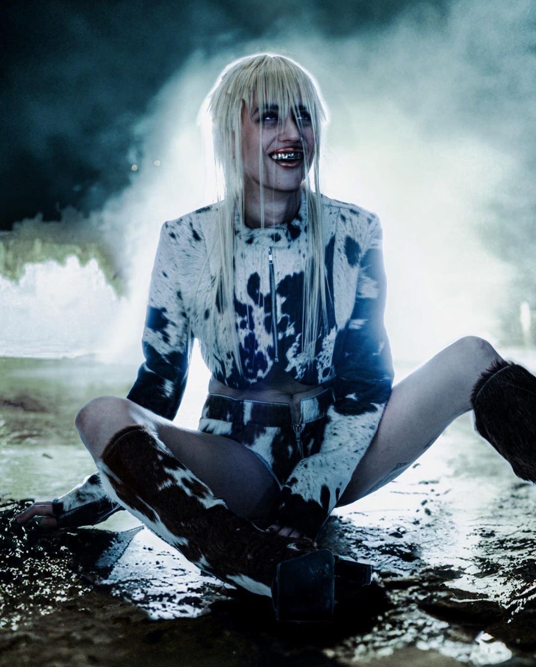 A woman with bleached blonde hair sits cross-legged, wearing a matching cow-print jacket, shorts, and boots in a moody, spotlight-lit setting.
