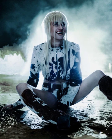 A woman with bleached blonde hair sits cross-legged, wearing a matching cow-print jacket, shorts, and boots in a moody, spotlight-lit setting.