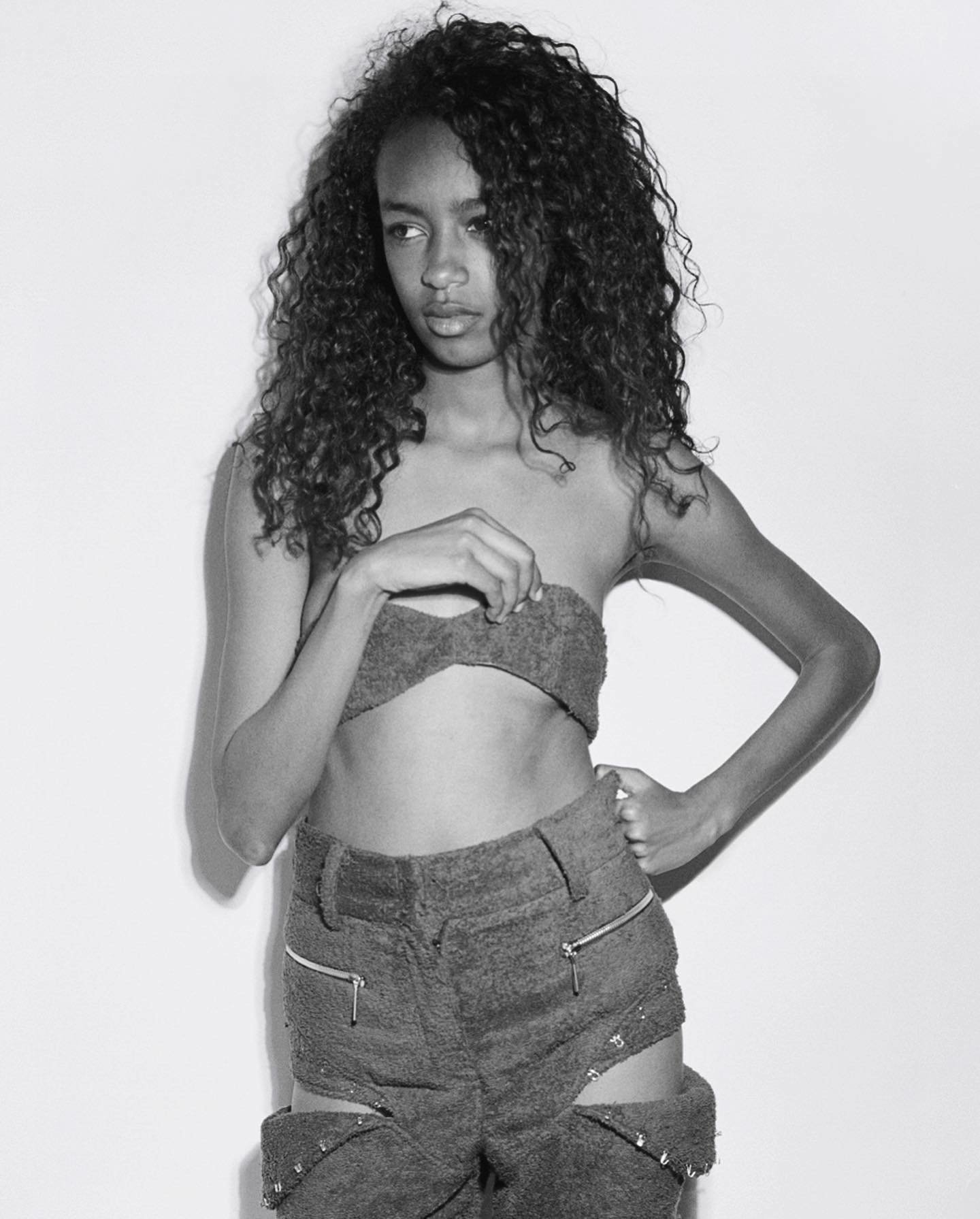 Portrait of a Black woman with curly hair wearing a knitted bra top and textured pants in a studio setting with a white background.