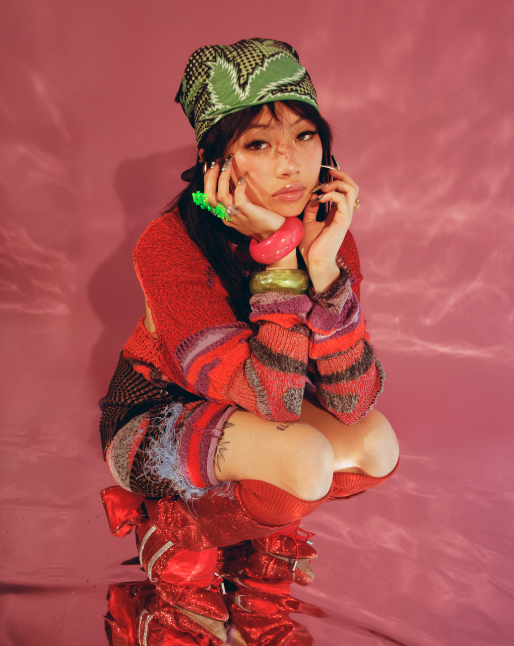 Asian woman crouching, wearing colorful knitwear and boots, gazing at the camera against a pink background, accessorized with bracelets and a bandana.