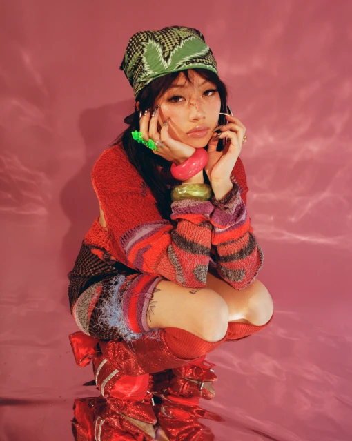 Asian woman crouching, wearing colorful knitwear and boots, gazing at the camera against a pink background, accessorized with bracelets and a bandana.