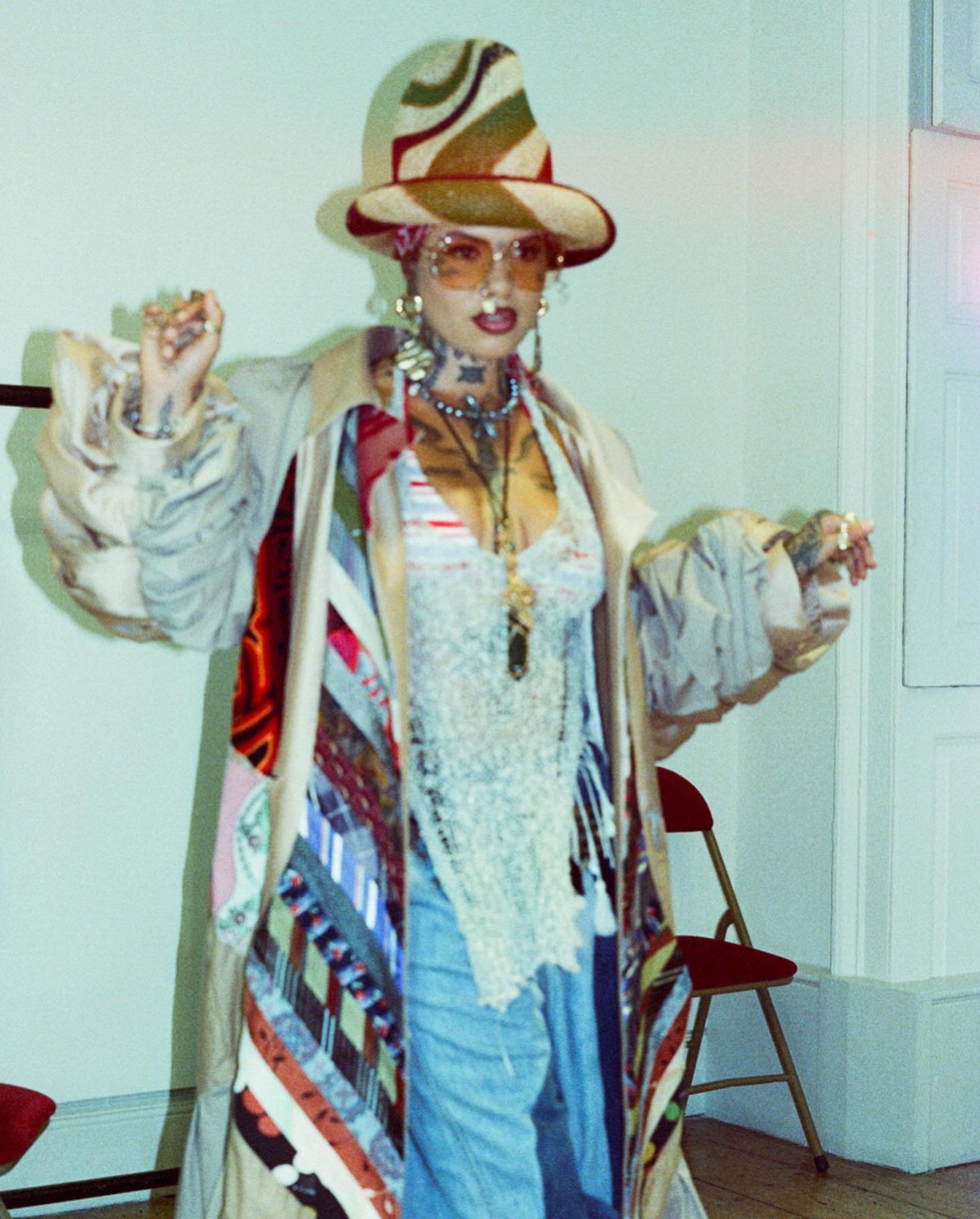 Adult female with stylish hat and coat stands indoors, wearing round sunglasses, multiple necklaces, a lace top, and distressed jeans.