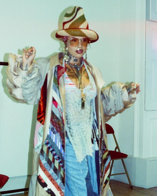 Adult female with stylish hat and coat stands indoors, wearing round sunglasses, multiple necklaces, a lace top, and distressed jeans.