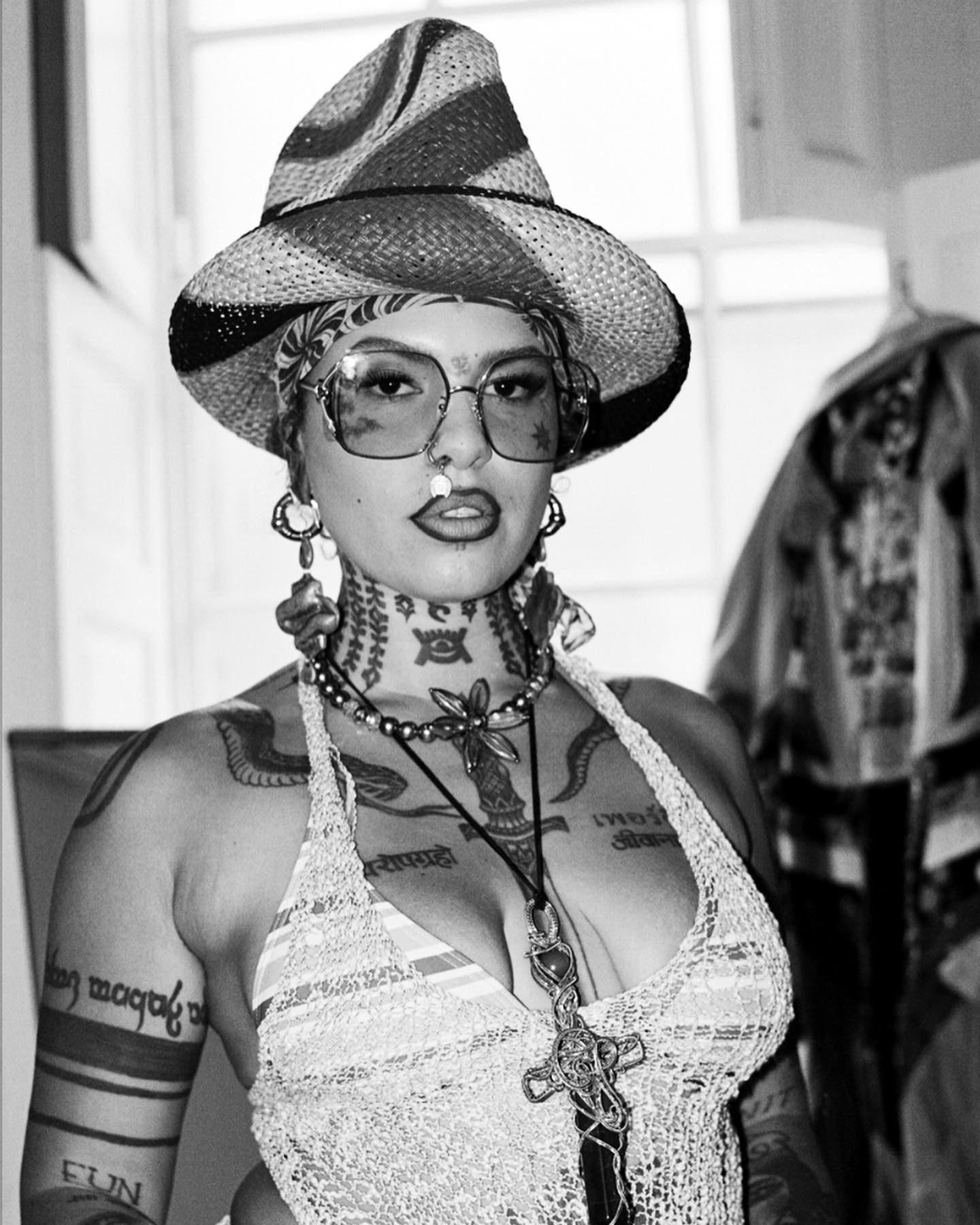 Portrait of an adult woman wearing a hat, glasses, and jewelry, with tattoos on her chest, neck, and arms in a black-and-white image.