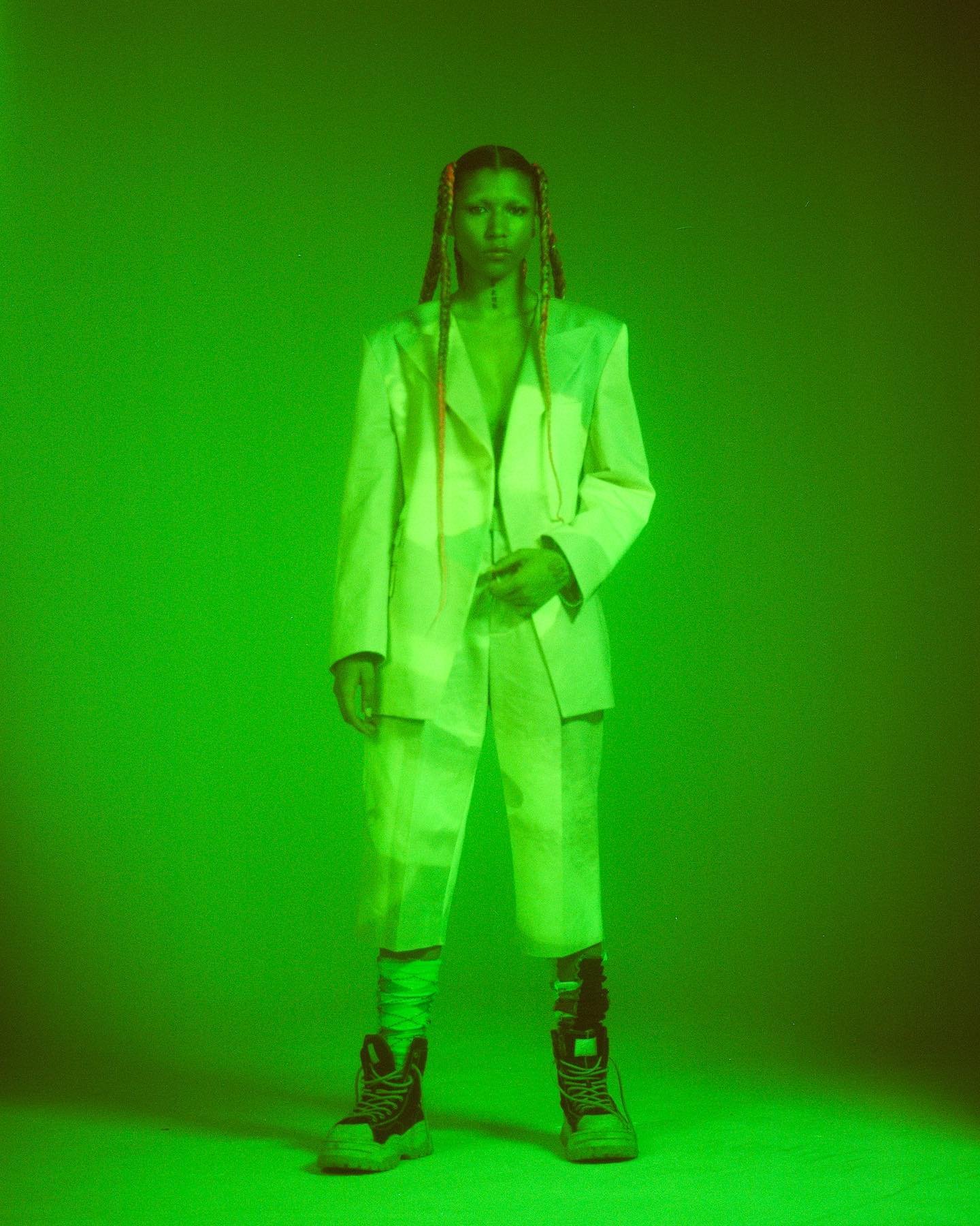 A standing adult woman in a light-colored suit and black boots under neon green lighting poses in front of a matching green background.