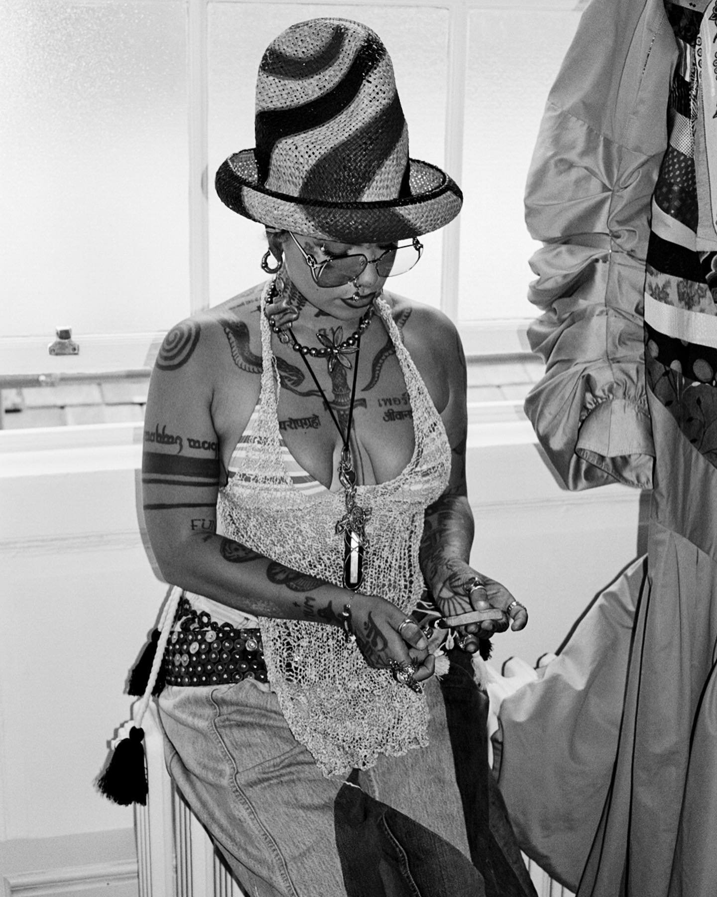 Black and white portrait of a tattooed adult female wearing a patterned hat, sheer halter top, and jeans, standing in an indoor environment.