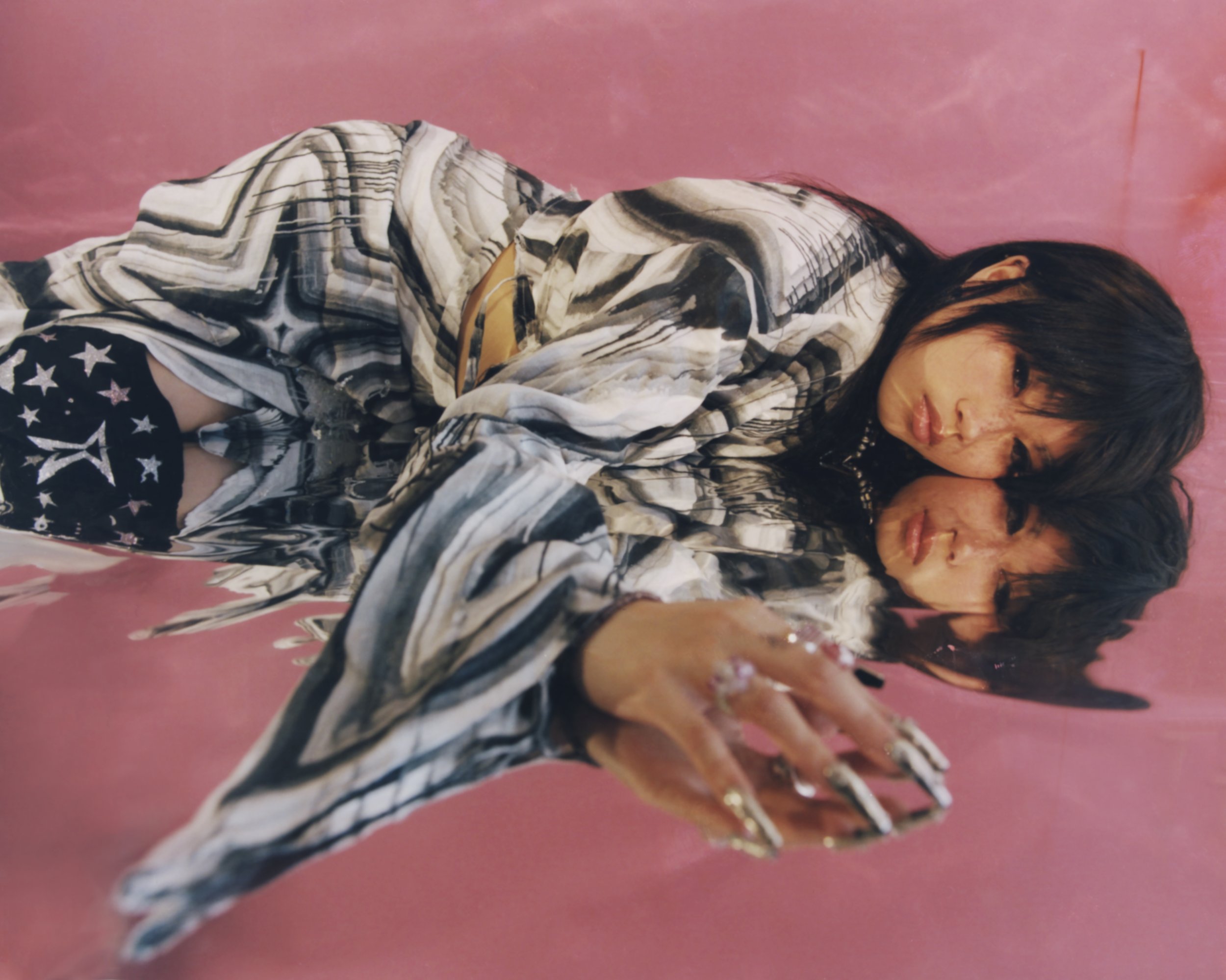 Young woman with long dark hair wearing patterned top and shorts lies on a reflective surface with a pink background and reflection visible.