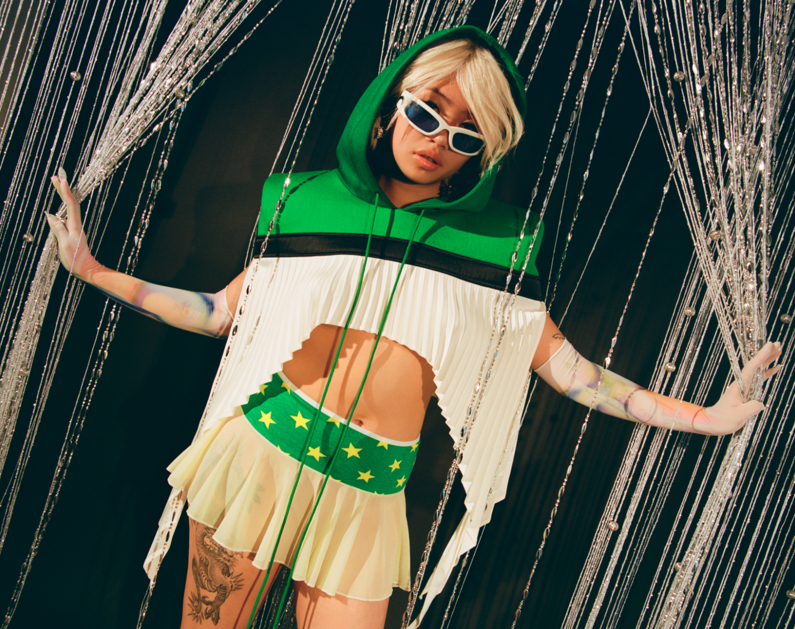 Woman poses in green and white hooded crop top and mini skirt against a string curtain and black background in a studio shot.