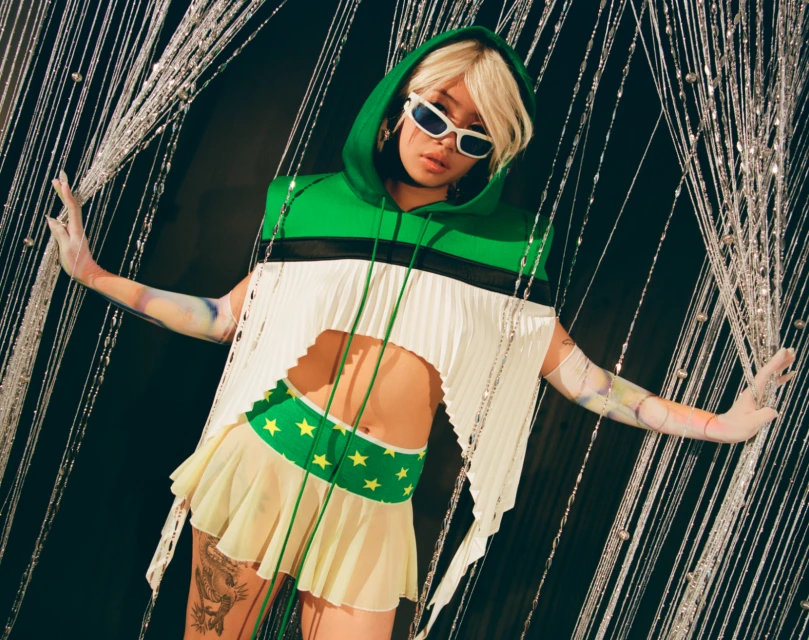 Woman poses in green and white hooded crop top and mini skirt against a string curtain and black background in a studio shot.