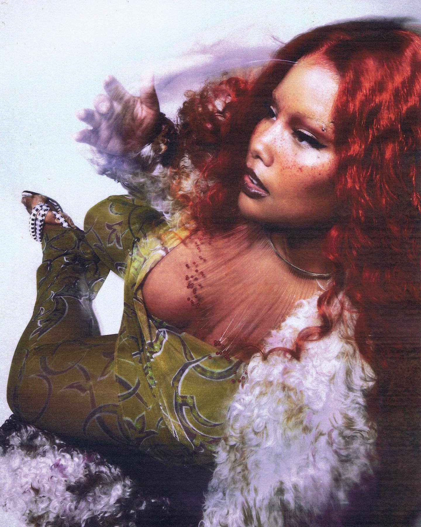 A fashion portrait features a woman with red hair, wearing a patterned olive jumpsuit with a faux fur collar and posing against a light background.