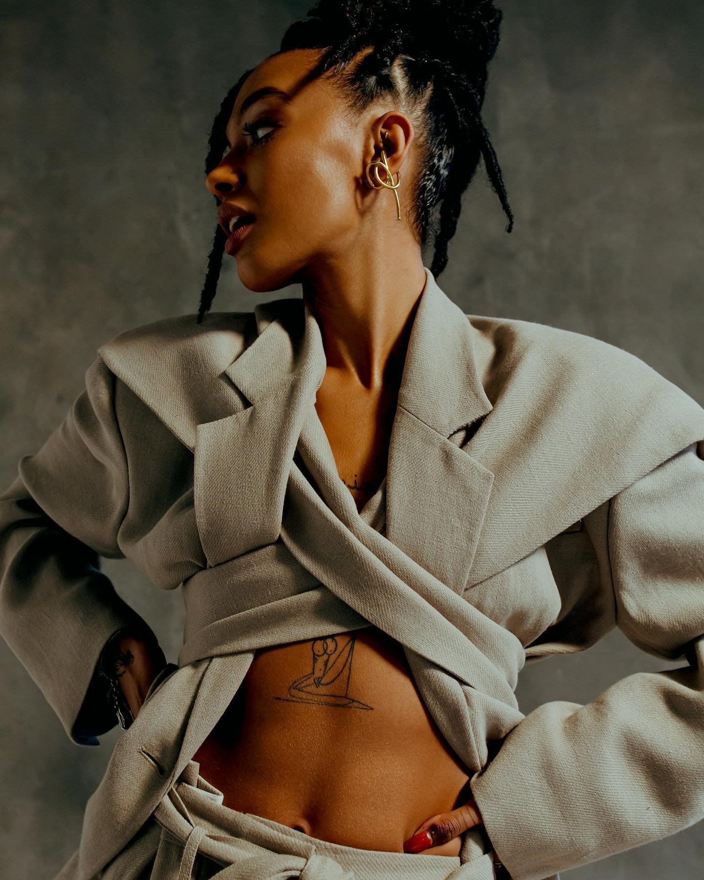 Portrait of a woman with braided hair and a beige wrap blazer, posing indoors against a grey background with soft lighting.