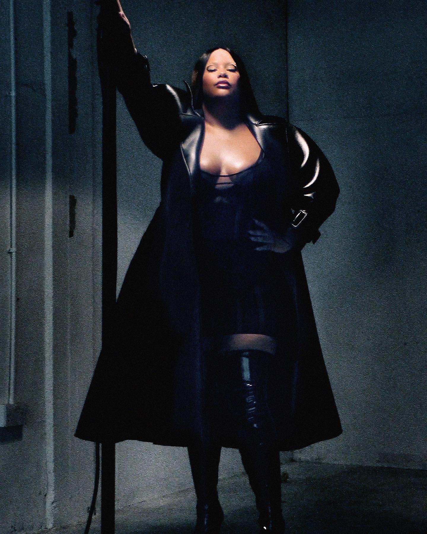 An adult female poses indoors wearing a black coat, dark lingerie, and thigh-high boots, standing near a wall with muted lighting.