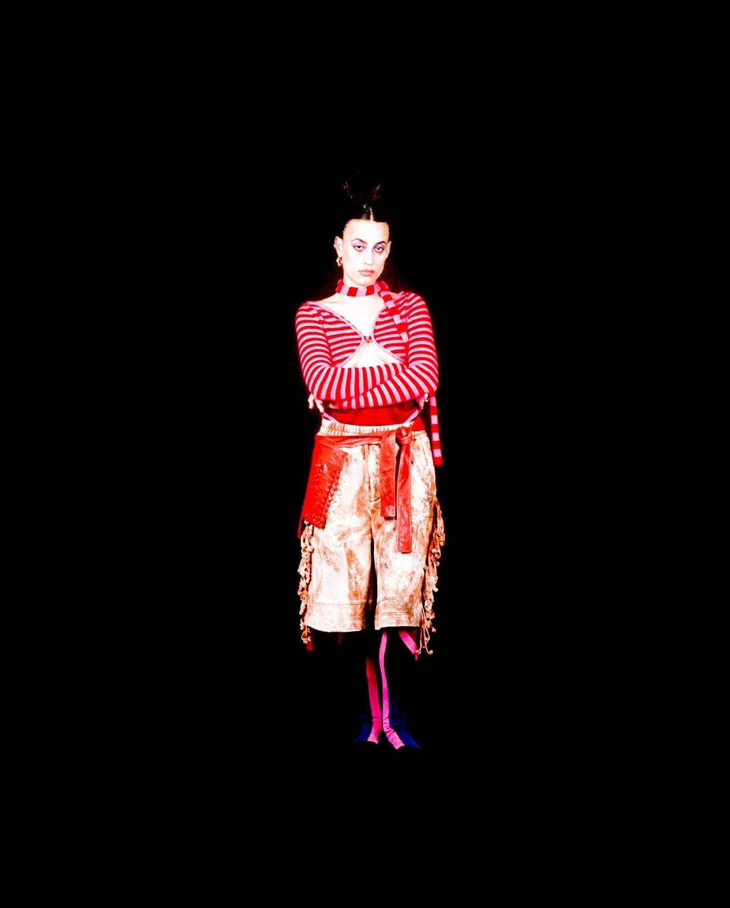 Full shot of a woman with crossed arms, wearing a striped top and shorts with a red belt against a black backdrop in a studio.