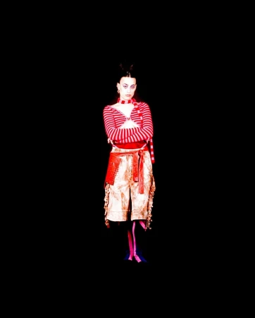 Full shot of a woman with crossed arms, wearing a striped top and shorts with a red belt against a black backdrop in a studio.