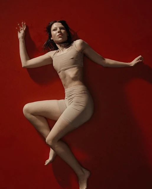 Adult female model lies on a red surface wearing a beige two-piece activewear set in a studio setting with soft, diffused lighting.