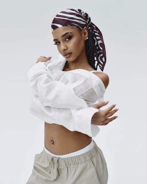 A fashionable young woman wearing a patterned headscarf, white blouse, and khaki pants strikes a casual pose against a neutral backdrop.
