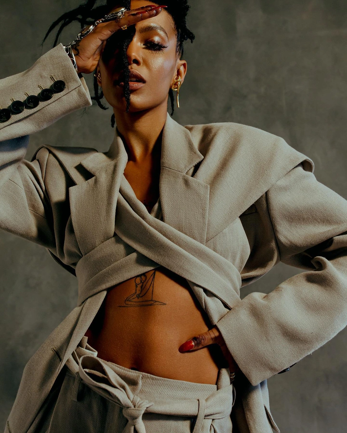 Close-up portrait of a woman wearing a structured beige belted jacket with crossed lapels against a dark textured wall background.