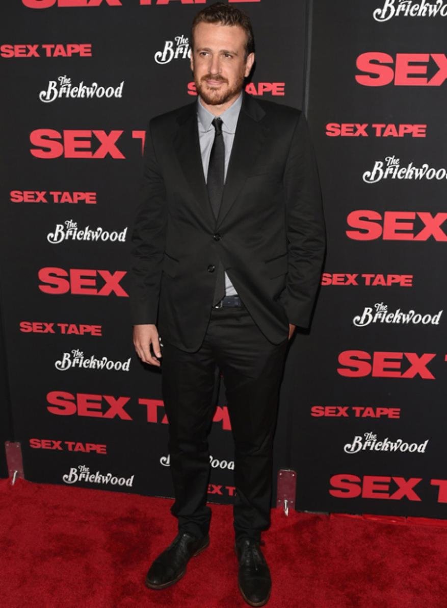Adult male with a beard stands in a black suit on a red carpet in front of a movie promo wall for 'Sex Tape' at a red-carpet event.
