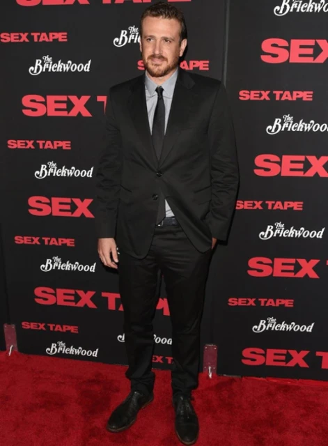 Adult male with a beard stands in a black suit on a red carpet in front of a movie promo wall for 'Sex Tape' at a red-carpet event.