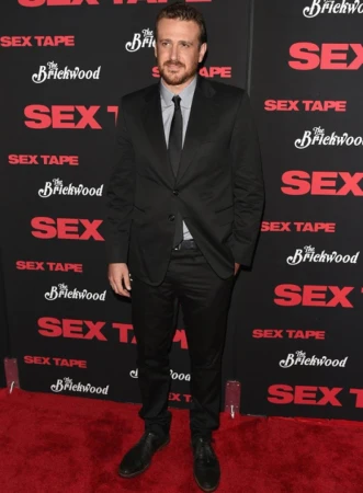Adult male with a beard stands in a black suit on a red carpet in front of a movie promo wall for 'Sex Tape' at a red-carpet event.