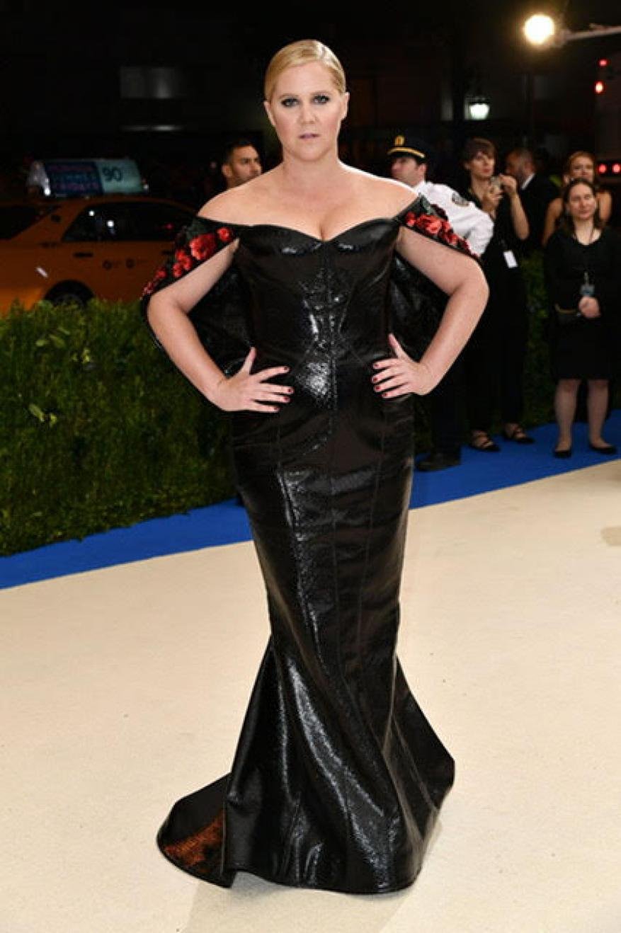 Amy Schumer at an event, wearing a black off-the-shoulder gown with red floral accents, posing with hands on hips.