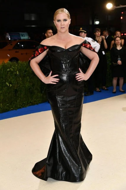 Amy Schumer at an event, wearing a black off-the-shoulder gown with red floral accents, posing with hands on hips.