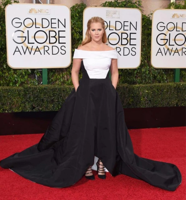 Amy Schumer poses in a white and black gown on the red carpet at the Golden Globe Awards event with a hedge background.