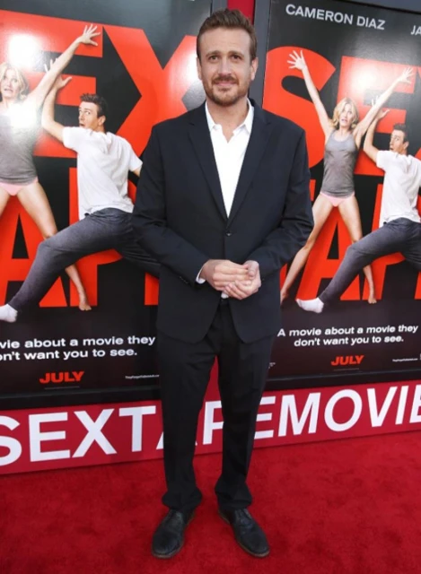 Jason Segel stands in a black suit at the 'Sex Tape' movie premiere on a red carpet in front of a movie poster backdrop.