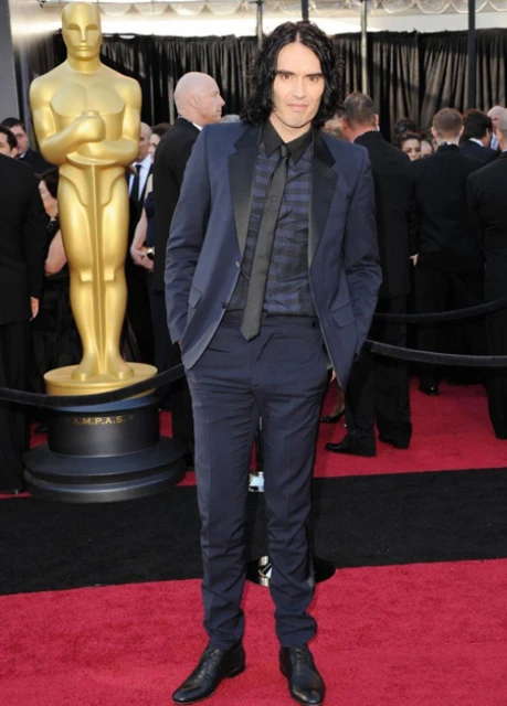 Russell Brand poses at an event standing on a red carpet in a blue suit, a dark shirt and a black tie. A golden statue is in the background.