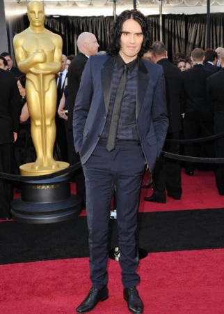 Russell Brand poses at an event standing on a red carpet in a blue suit, a dark shirt and a black tie. A golden statue is in the background.