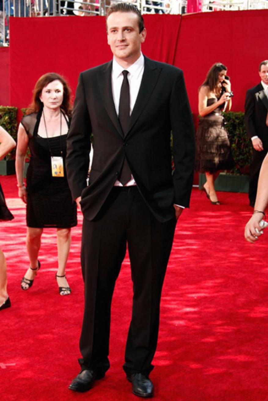 Jason Segel wearing a black suit and tie standing on a red carpet at an event