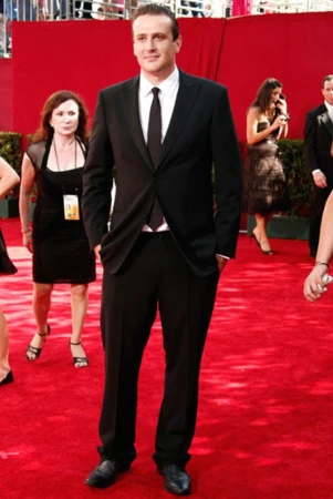 Jason Segel wearing a black suit and tie standing on a red carpet at an event