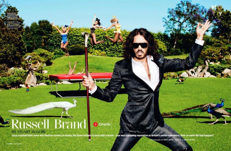 Russell Brand poses outdoors in a suit with bunny dancers jumping in the background, peacocks and a daybed also visible.