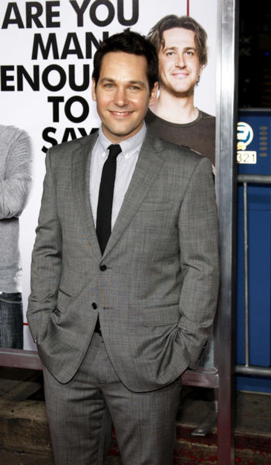 Paul Rudd poses in a grey suit with a black tie on a red carpet in front of a movie poster featuring another person's portrait.