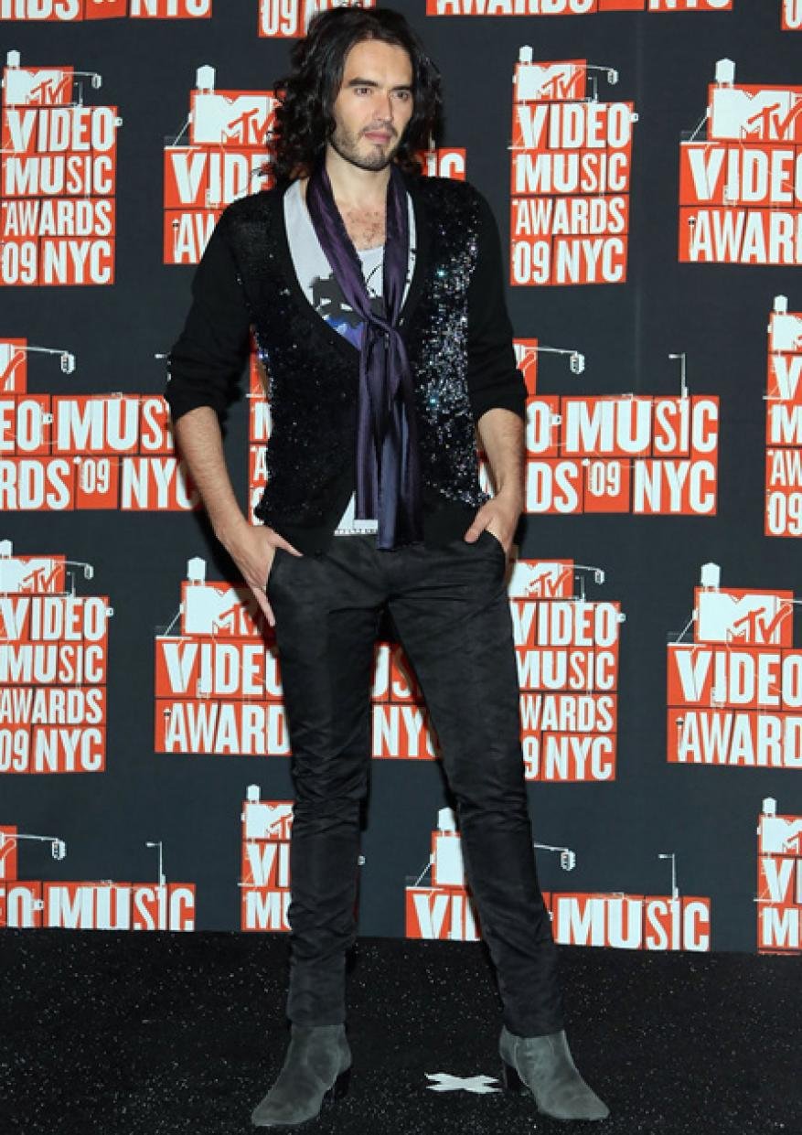 Russell Brand poses in front of MTV Video Music Awards backdrop wearing a patterned sweater, dark pants, and heeled boots at event.