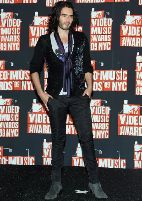 Russell Brand poses in front of MTV Video Music Awards backdrop wearing a patterned sweater, dark pants, and heeled boots at event.
