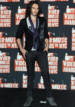 Russell Brand poses in front of MTV Video Music Awards backdrop wearing a patterned sweater, dark pants, and heeled boots at event.