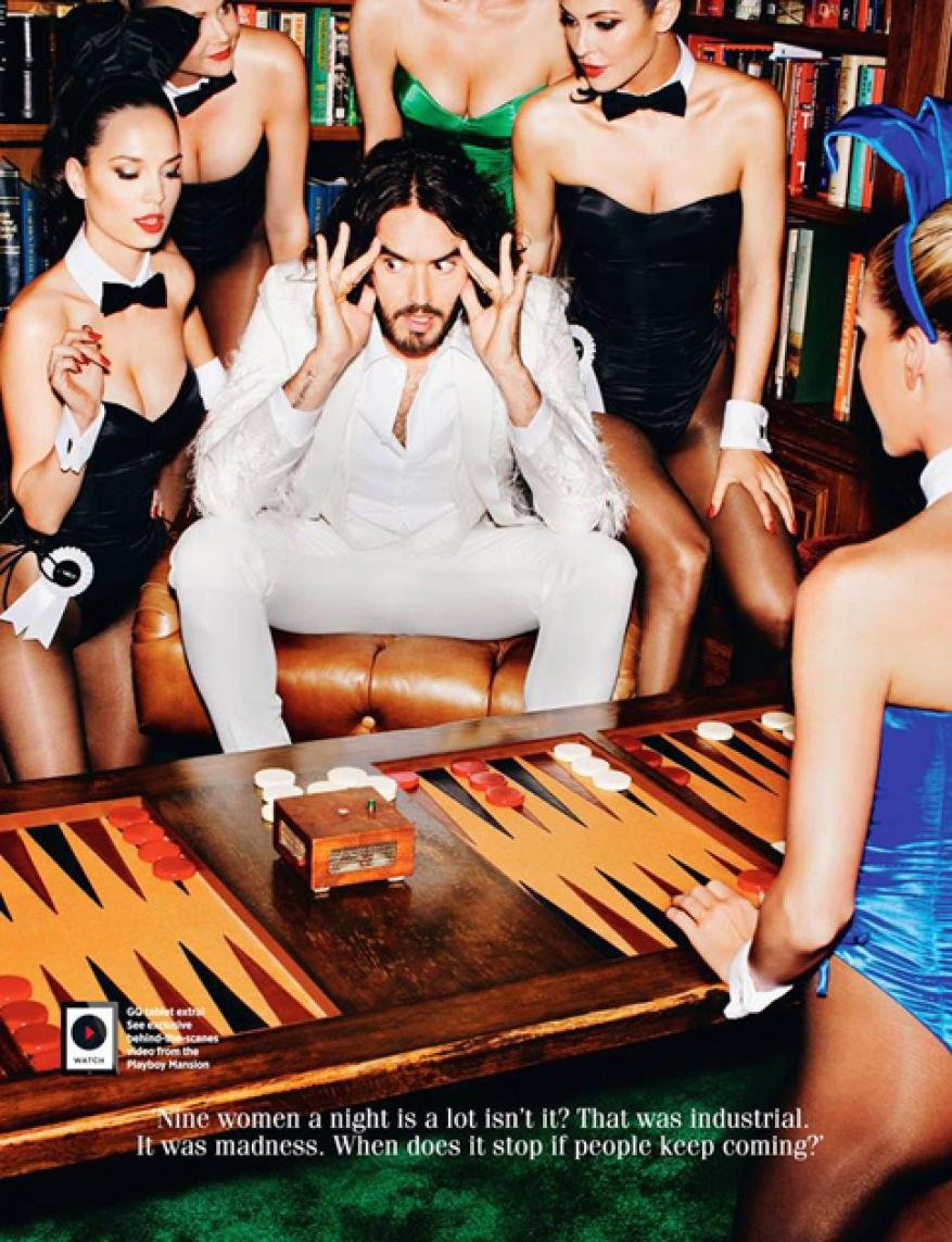Russell Brand poses with women in bunny costumes at a backgammon table in a room lined with books, creating a vintage, playful image.