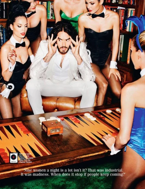 Russell Brand poses with women in bunny costumes at a backgammon table in a room lined with books, creating a vintage, playful image.